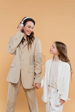 happy mother and daughter in suits, woman wearing wireless headphones near girl and holding hands on beige background, fashionable outfits, formal attire, corporate mom, modern family 