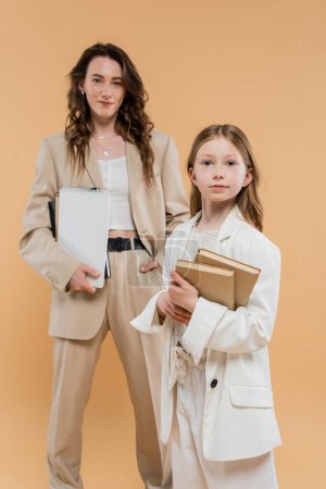trendy mother and daughter in suits, child holding books near blurred woman with notebooks on beige background, fashionable outfits, formal attire, corporate mom, education concept 