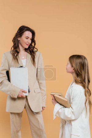fashionable family, mother and daughter in suits, woman and child holding books and notebooks while standing together on beige background, formal attire, corporate mom, education concept 