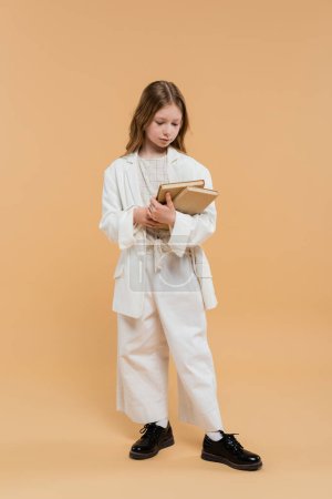 education concept, preteen girl in white suit holding books and standing on beige background, fashionable outfit, formal attire, back to school, preparing for school new year 