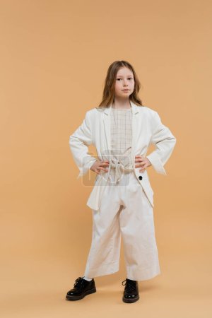 trendy preteen girl in white suit and black shoes looking at camera while standing on beige background, fashionable outfit, formal attire, child model, trendsetter, style, posing with hands on hips
