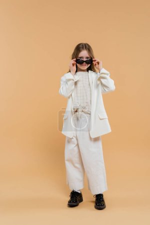trendy preteen girl in white suit and black shoes looking at camera while wearing sunglasses and standing on beige background, fashionable outfit, formal attire, child model, trendsetter, style 