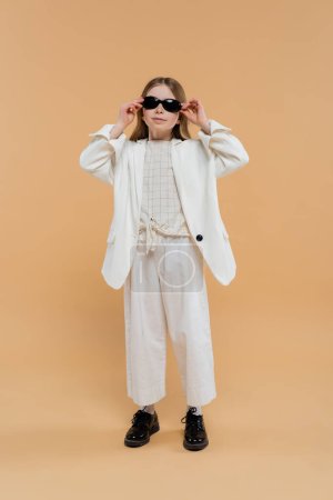 stylish preteen girl in white suit and black shoes looking at camera while wearing sunglasses and standing on beige background, fashionable outfit, formal attire, child model, trendsetter, style 