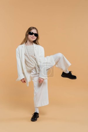 trendy preteen girl in white suit, sunglasses and black shoes posing with raised leg and standing on beige background, fashionable outfit, formal attire, child model, trendsetter, style 