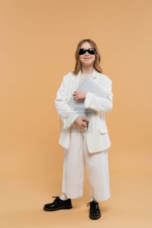 online education, trendy preteen girl in white suit, happy kid in sunglasses holding laptop and standing on beige background, fashionable outfit, formal attire, child model, trendsetter, e-learning 