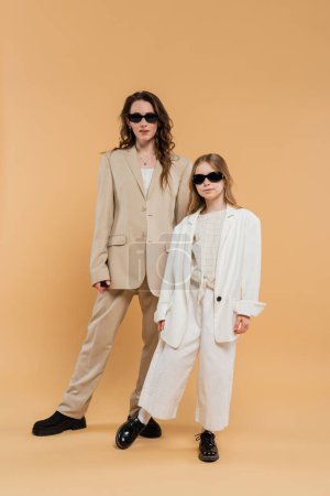 madre e hija de moda en gafas de sol, mujer de negocios y chica en trajes de pie juntos sobre fondo beige, trajes de moda, atuendo formal, mamá corporativa, familia moderna 