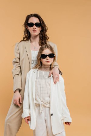 moderne Familie, stilvolle Mutter und Tochter mit Sonnenbrille, Geschäftsfrau und Mädchen in Anzügen, die zusammen auf beigem Hintergrund stehen, modische Outfits, formelle Kleidung, Corporate Mom 