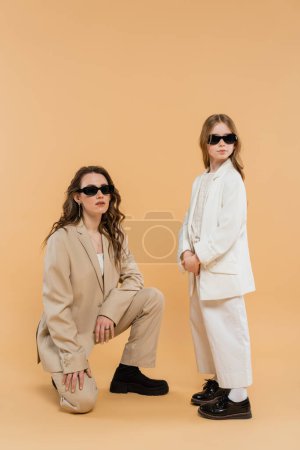 modern family, stylish mother and daughter in suits and sunglasses, businesswoman sitting near girl on beige background, fashionable outfits, formal attire, corporate mom 