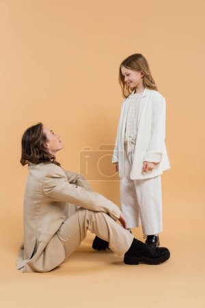 mother child bonding concept, stylish woman in suit sitting and looking at happy preteen daughter on beige background, corporate mom, businesswoman, motherly love, smiling girl 