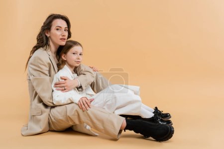 Mutter-Kind-Bindungskonzept, stilvolle Frau im Anzug umarmt preteen Tochter und steht zusammen in Anzügen auf beigem Hintergrund, korporative Mutter, Geschäftsfrau, mütterliche Liebe 
