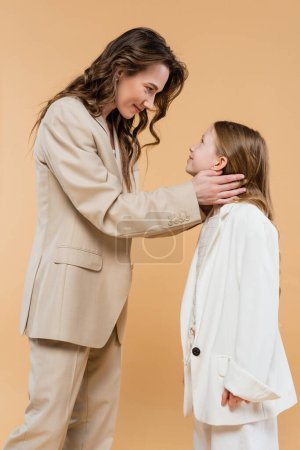 mother child bonding concept, happy woman in suit hugging cheerful preteen daughter and standing together in suits on beige background, corporate mom, businesswoman, motherly love 