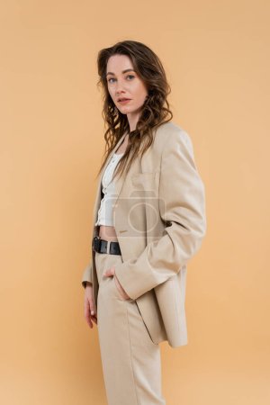 style and fashion concept, young woman with wavy hair standing in fashionable suit while posing on beige background, formal attire, hand in pocket, looking at camera, modern elegance 
