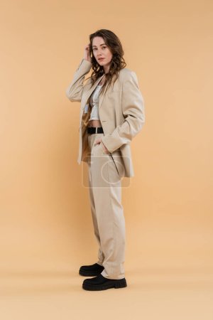 style et concept de mode, jeune femme aux cheveux ondulés debout en costume à la mode et regardant la caméra tout en posant sur fond beige, main dans la poche, élégance moderne