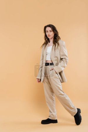 fashion trend concept, young woman with wavy hair walking in fashionable suit and looking at camera on beige background, hand in pocket, classic style, professional attire 