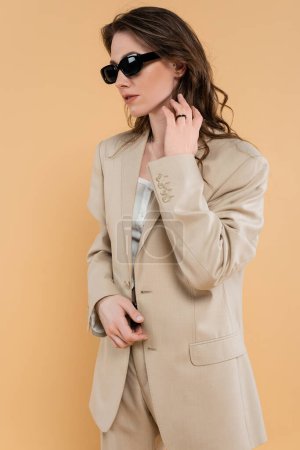 fashion trend concept, young woman with wavy hair standing in fashionable suit and sunglasses on beige background, classic style, chic stylish posing, professional attire, formal attire 