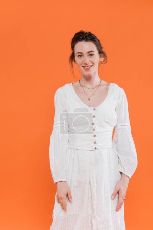 mode d'été, jeune femme heureuse en robe de soleil blanche souriante et regardant la caméra sur fond orange, fond vibrant, posant élégant, dame en blanc 