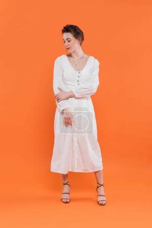 Foto de Moda de verano, mujer joven en vestido de sol blanco mirando hacia otro lado y de pie sobre fondo naranja, fondo vibrante, posando con estilo, señora de blanco, tendencia de la moda, elegancia - Imagen libre de derechos