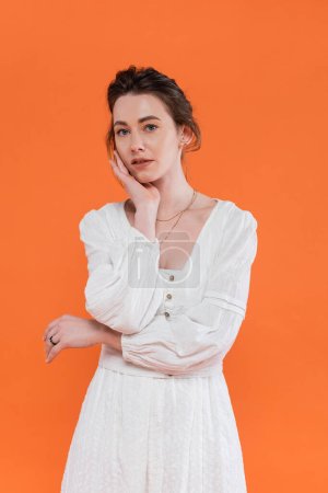 tendencias de verano, mujer joven en vestido de sol blanco mirando a la cámara y de pie sobre fondo naranja, fondo vibrante, posando con estilo, dama de blanco, modelo de moda, elegancia 