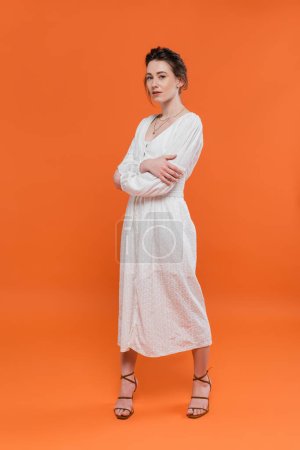 summer trends, model in white sun dress looking at camera and standing with folded arms on orange background, stylish posing, lady in white, vibrant background, fashion, young woman 