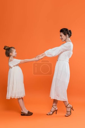 summer trends, happy young mother holding hands with preteen daughter and standing on orange background, white sun dresses, togetherness, fashion and style concept, bonding and love 