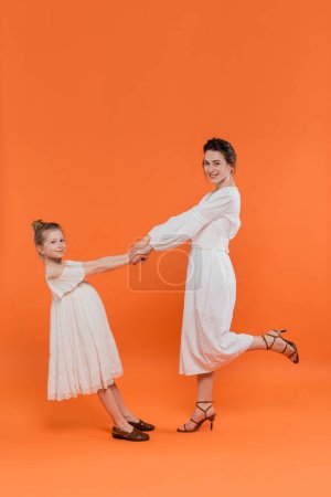 tendencias de verano, madre joven cogida de la mano con hija preadolescente y de pie sobre fondo naranja, vestidos de sol blanco, unión, moda y concepto de estilo, vinculación, alegre 