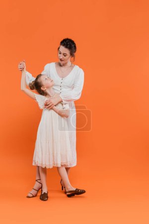 tendencias de verano, madre cogida de la mano con hija preadolescente y de pie juntos sobre fondo naranja, vestidos de sol blanco, unión, moda y concepto de estilo, vinculación y amor 