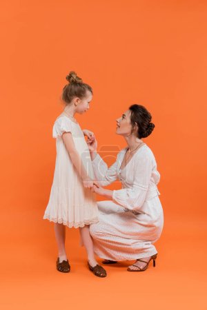 Sommertrends, staunende junge Mutter hält Händchen mit frühgeborener Tochter auf orangefarbenem Hintergrund, weiße Sonnenkleider, Zweisamkeit, Mode- und Stilkonzept, Bindung und Liebe 
