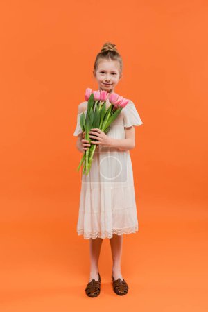 Sommermode, glückliches Mädchen im weißen Sonnenkleid mit rosa Tulpen auf orangefarbenem Hintergrund, Mode- und Stilkonzept, Blumenstrauß, modisches Kind, lebendige Farben, volle Länge 