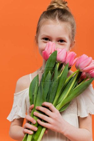 Téléchargez les photos : Fille préadolescente en robe de soleil blanche tenant des tulipes roses sur fond orange, concept de mode et de style, bouquet de fleurs, enfant à la mode, couleurs vives, couvrant le visage - en image libre de droit
