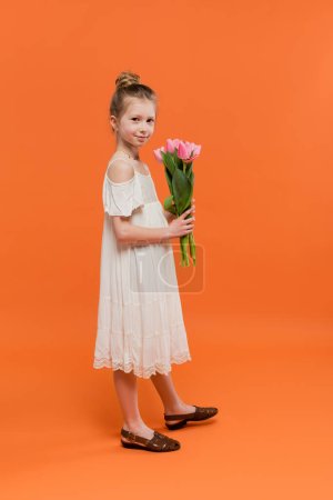 Téléchargez les photos : Robe d'été, preteen girl en robe de soleil blanche tenant des tulipes roses sur fond orange, concept de mode et de style, bouquet de fleurs, enfant à la mode, couleurs vives, robe d'été, pleine longueur - en image libre de droit