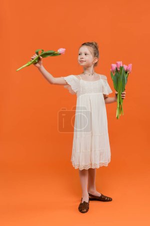 Foto de Ramo de flores, niña preadolescente en vestido de sol blanco con tulipanes rosados sobre fondo naranja, concepto de moda y estilo, niño de moda, colores vibrantes, moda de verano, niño lindo - Imagen libre de derechos