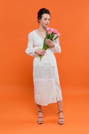 Téléchargez les photos : Bouquet de fleurs, jeune femme en robe de soleil blanche tenant des tulipes et debout sur fond orange, pose élégante, dame en blanc, fond vibrant, mode, été, pleine longueur - en image libre de droit