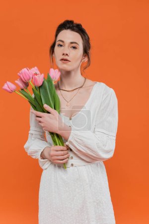 Téléchargez les photos : Bouquet de fleurs, jolie jeune femme en robe d'été blanche tenant des tulipes et debout sur fond orange, pose élégante, dame en blanc, fond vibrant, mode, été - en image libre de droit