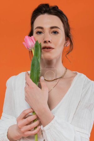moda cotidiana, mujer joven en vestido de sol blanco sosteniendo tulipán rosa y de pie sobre fondo naranja, dama en blanco, fondo vibrante, moda y naturaleza, verano, retrato 