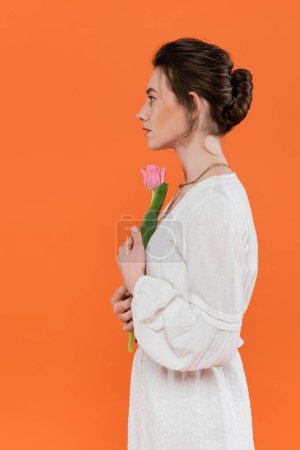 Foto de Vista lateral de mujer joven en vestido de sol blanco sosteniendo tulipán rosa y de pie sobre fondo naranja, dama en blanco, fondo vibrante, moda, verano, retrato, sensualidad, moda cotidiana - Imagen libre de derechos