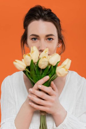 Téléchargez les photos : Bouquet de fleurs, mode quotidienne, jeune femme en robe de soleil blanche tenant des tulipes jaunes et debout sur fond orange, dame en blanc, fond vibrant, mode, été - en image libre de droit