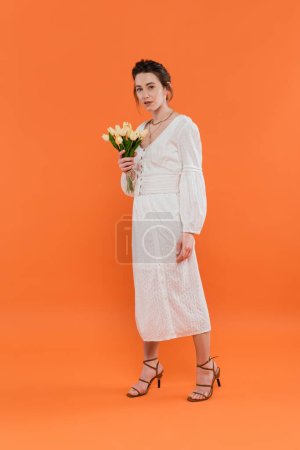concepto de moda, mujer bastante joven en vestido de sol blanco sosteniendo tulipanes amarillos y de pie sobre fondo naranja, dama en blanco, fondo vibrante, moda, verano, ramo de flores, longitud completa