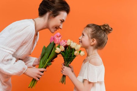 feliz madre e hijo con flores, mujer joven y niña sosteniendo tulipanes y mirándose el uno al otro sobre fondo naranja, moda de verano, vestidos de sol, vinculación femenina 