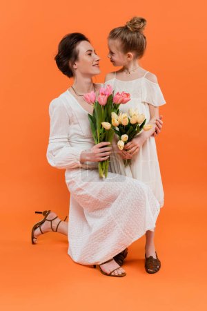 feliz madre e hija con flores, mujer joven y niña sosteniendo tulipanes y posando sobre fondo naranja, moda de verano, vestidos de sol, vinculación femenina 
