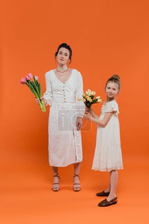 Foto de Feliz madre e hija con tulipanes, mujer joven y niña sosteniendo flores y posando sobre fondo naranja, moda de verano, vestidos de sol, vinculación femenina, longitud completa, ocasión especial - Imagen libre de derechos