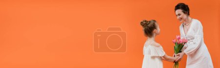 Mother`s day, preteen girl giving bouquet of flowers to smiling mother on orange background, bonding, white dresses, pink tulips, happy holiday, vibrant colors, joyful occasion, banner 