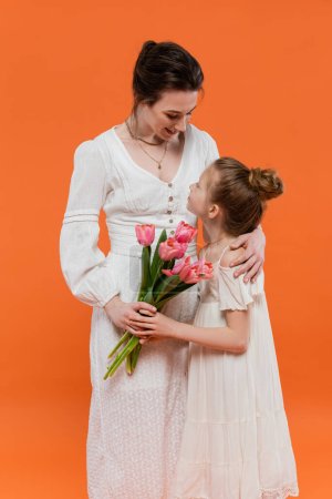 Mother`s day, mother hugging preteen daughter with bouquet of flowers on orange background, bonding, white dresses, pink tulips, happy holiday, vibrant colors, joyful occasion 