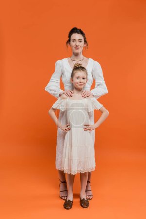 Foto de Crianza moderna, moda de verano, mujer abrazando hija preadolescente y de pie juntos en vestidos de sol blanco sobre fondo naranja, vinculación femenina, familia de moda, estilo de verano - Imagen libre de derechos