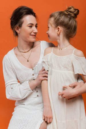 Mutterliebe, hübsche Mutter lächelnd und Tochter auf orangefarbenem Hintergrund betrachtend, weiße Sonnenkleider, Sommermode, Zweisamkeit, Liebe, weibliche Bindung, Frauenstil, moderne Elternschaft 