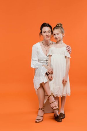 motherly love, brunette young woman sitting on chair and hugging preteen daughter on orange background, white sun dresses, summer fashion, togetherness, love, female bonding, modern parenting 