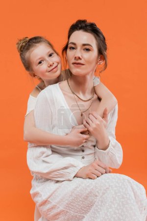 Mutter und Tochter, glückliches, junges Mädchen umarmt junge Frau auf orangefarbenem Hintergrund, weiße Sonnenkleider, moderne Elternschaft, Sommermode, Zweisamkeit, Liebe, modische Familie 