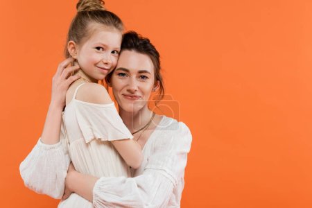 family bonding, joyful mother and daughter hugging each other on orange background smile, white sun dresses, modern parenting, summer fashion, togetherness, mom and her child 