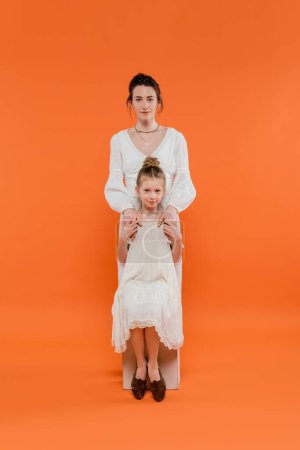 Mutterliebe, junge Mutter umarmt Tochter und steht zusammen in weißen Sonnenkleidern auf orangefarbenem Hintergrund, Familienmode, modische Frau und Mädchen, weibliche Bindung, moderne Elternschaft 