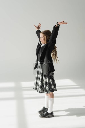 Téléchargez les photos : Écolière en uniforme, joyeux preteen girl debout avec les mains levées sur fond gris, tenue formelle, enfant à la mode, joyeux, excitation, célébration de l'apprentissage, concept de retour à l'école - en image libre de droit