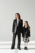 working mother and schoolgirl, cheerful girl in school uniform standing with businesswoman in suit on grey background, holding hands, formal attire, fashionable family, bonding, modern parenting  Poster #659560034